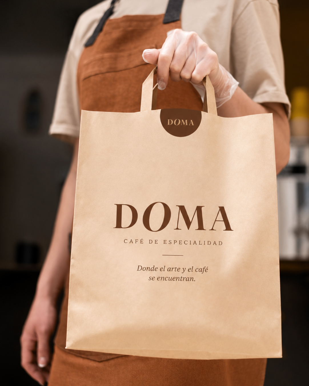 Bolsa kraft de DOMA con logotipo y lema, sostenida por una barista